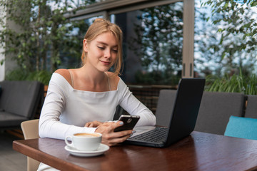 Modern business woman is reading incoming sms message on smartphone or watching broadcasting online on modern mobile phone while sitting in caffe with a laptop. Female student sitting in coffee.