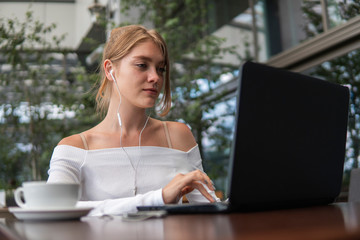 Beautiful young woman in white t-shirt is working on laptop and smiling while sitting outdoors in cafe. Young female using laptop for work. Female freelancer working on laptop in an outdoor cafe.