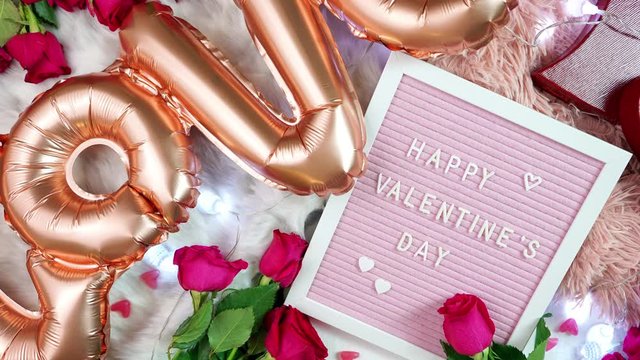 Happy Valentine's Day flat lay overhead with roses, chocolate gift box and letterboard with greeting text and large rose gold balloons in shape of the word Love. Slow panning shot.