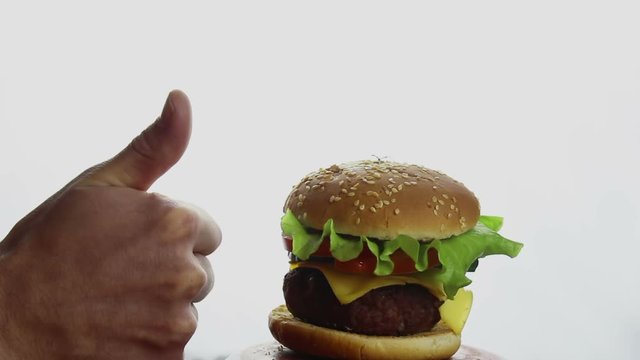 Male hand shows thumb up on fresh juicy burger. Large juicy burger with beef cutlet, fresh vegetables and melted cheese. Fast food, high-calorie food.Burger Close-up on a white background.
