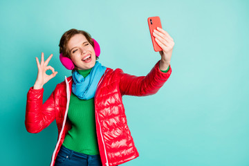 Alright. Photo of funny pretty lady hold telephone making selfies winking eye showing okay symbol wear red coat scarf pink ear muffs jumper pants isolated teal color background © deagreez