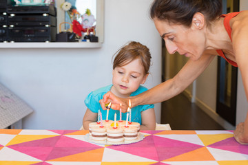 Obraz na płótnie Canvas woman lighting candles with lighter in hand on birthday cake on colorful tablecloth at home, and three years old blonde child looking