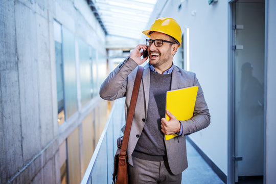 Handsome smiling positive hardworking architect holding folder with important documents and talking on the phone with constructor.