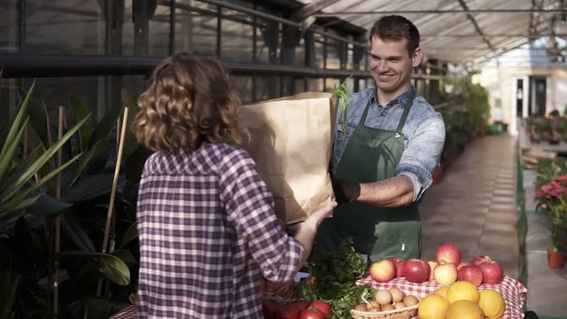 Potrait of pleased, smiling female customer recieving a paper bag with organic grocery. Caucasian,tall salesman in green apron is selling vegetables in indoors greenhouse