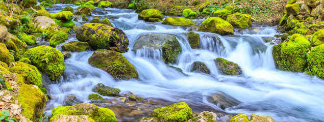 A small stream with a waterfall and mossy rocks in spring, panoramic image