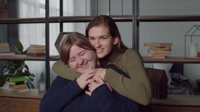 Lifestyle portrait of happy attractive young deaf women embracing in domestic interior, looking with beaming genuine smiles, showing i love you using sign language, expressing happiness and joy.
