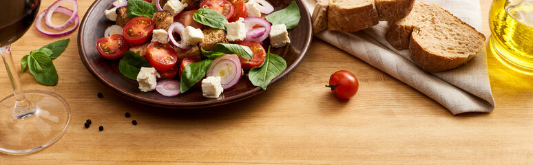 delicious Italian vegetable salad panzanella served on plate on wooden table near fresh ingredients, bread and red wine, panoramic shot