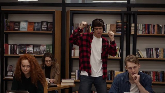 Joyful excited handsome european man in checkered shirt performing expressive dance while listening groovy music in headphones in academic library against bookshelves background and classmates around