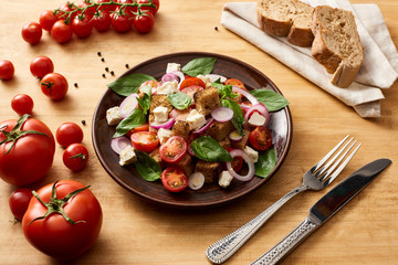 delicious Italian vegetable salad panzanella served on plate on wooden table near fresh tomatoes, bread, fork and knife