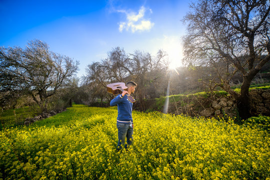 Joven m&uacute;sico con una guitarra en el hombro, buscando un lugar para sentarse y tocar en un campo de flores amarillas.
