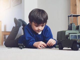 Portrait of School boy lying down on the carpet floor playing with soldiers, military car and figurine toys, Happy Kid playing wars and peace on his own at home, Children imagination and development