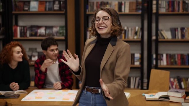 Portrait of joyful excited european woman in jacket and jeans performing expressive dance while listening music in headphones in academic library against bookshelves background and classmates around