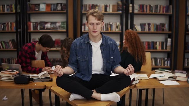 In the library during the studying. Young male student meditating on the desk in the lotos pose while his classmates on the background continuing studying ignoring him. Bookshelves on the background