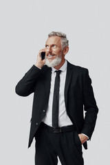 Happy mature man in full suit talking on the phone and smiling while standing against grey background