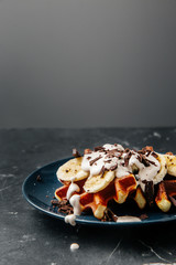 Viennese waffles on a plate, with a crumb of chocolate and slices of banana on a dark textured background.