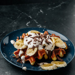 Viennese waffles on a plate, with a crumb of chocolate and slices of banana on a dark textured background.