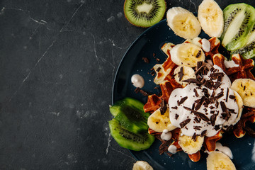 Viennese waffles on a plate, with cream sprinkled with a crumb of chocolate, slices of banana and kiwi on a dark textured background.
