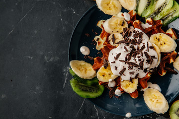 Viennese waffles on a plate, with cream sprinkled with a crumb of chocolate, slices of banana and kiwi on a dark textured background.