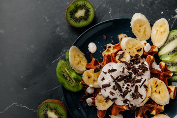 Viennese waffles on a plate, with cream sprinkled with a crumb of chocolate, slices of banana and kiwi on a dark textured background.