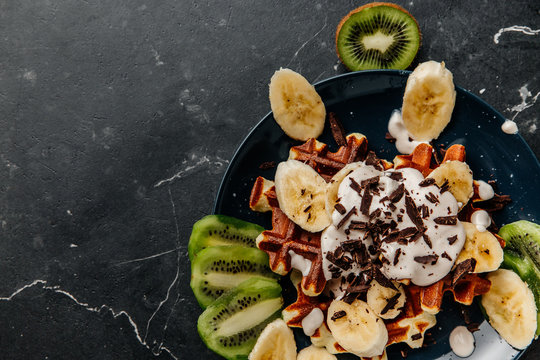 Viennese waffles on a plate, with cream sprinkled with a crumb of chocolate, slices of banana and kiwi on a dark textured background. - Powered by Adobe