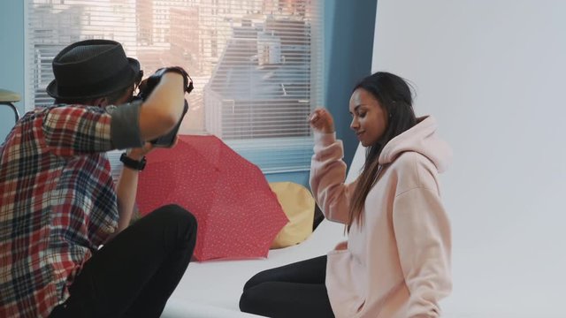 Close-up of make-up artist applying makeup on beautiful black model sitting on the floor and posing. Photographer taking photos on professional camera.