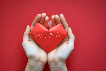 Women's hands hold a red heart with a cardiogram made of origami paper on a red background. Valentine's day card. Space for text.