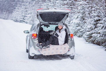 Woman sits in trunk of the car and holds a cup of hot tea in hands. Winter vacation, travel. Snow forest and road. Snowy weather