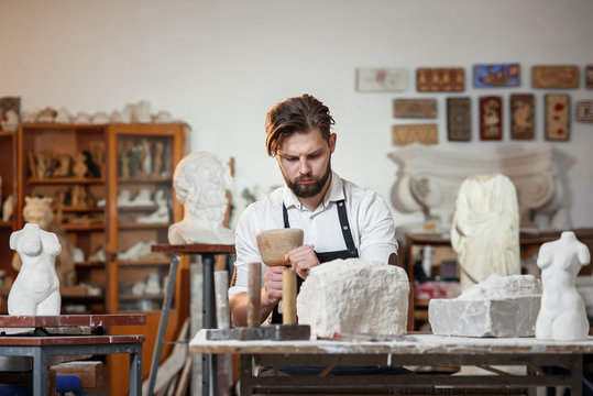 Bearded craftsman works in white stone carving with a chisel. Creative workshop with works of art.