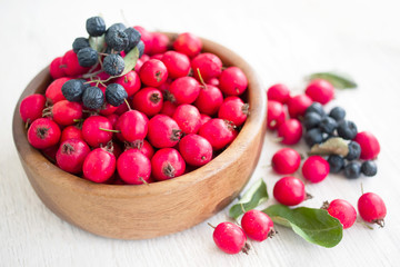Fresh red berries of hawthorn and chokeberry in a wooden round bowl on a white table surface, close up