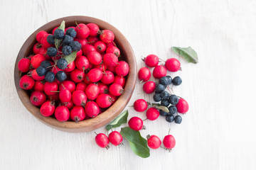 Fresh red berries of hawthorn and chokeberry in a wooden round bowl on a white table surface, close up