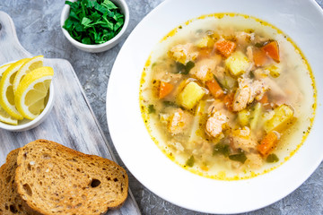 Fish soup made of salmon and vegetables in white bowl, served with bread toasts, parsley, lemon on textured grey table. Vegetarian and healthy food rich with Omega 3. Flat lay with close up view.