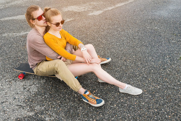 An attractive young couple in sunglasses sit back to back on their longboard in a suburban parking. The concept of a young family generation millennials style and leisure