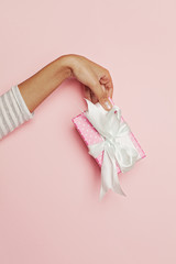 Woman hand holding gift box with white silky ribbon and bow on pink background with copy space