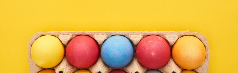 top view of multicolored painted Easter eggs in cardboard container on yellow background, panoramic shot