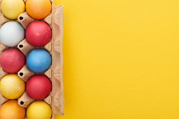 top view of multicolored painted Easter eggs in cardboard container on yellow background with copy space