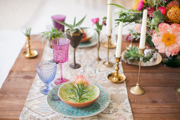Served table for a wedding dinner in boho chic style. Decor from fresh flowers of peonies, roses, orchids and Leucospermum conocarpodendron. Crockery plates, glasses and candles. Wooden table.