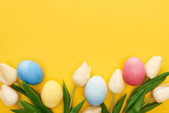 Top view of tulips and painted Easter eggs on colorful yellow background
