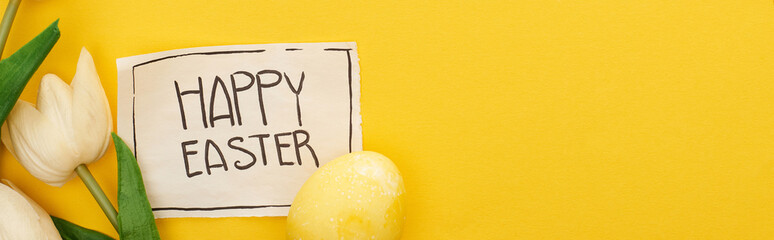 top view of Easter egg, tulips and greeting card with happy Easter lettering on yellow colorful background, panoramic shot