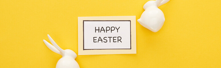 top view of greeting card with happy Easter lettering near Easter white bunnies on yellow colorful background, panoramic shot