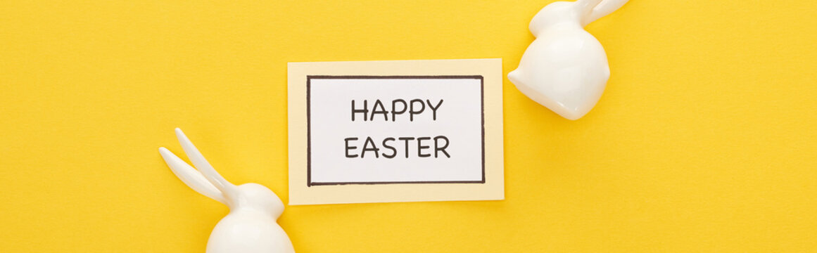 top view of greeting card with happy Easter lettering near Easter white bunnies on yellow colorful background, panoramic shot