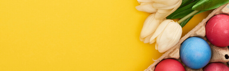 Top view of tulips and multicolored painted Easter eggs in cardboard container on colorful yellow background, panoramic shot