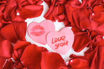 Two heart shaped stickers with a love note and womans girl red lipstick kiss mark and red rose petals on the white wooden table. Attributes for the celebration of Valentine's day