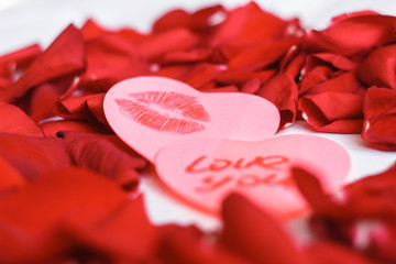Two heart shaped stickers with a love note and womans girl red lipstick kiss mark and red rose petals on the white wooden table. Attributes for the celebration of Valentine's day. Close-up