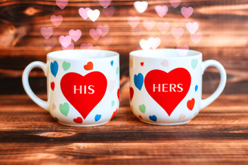 Two thematic white mugs with hearts on a background of wooden boards.  Attributes for the celebration of Valentine's day. A mug for him and a mug for her with bokeh in the shape of hearts