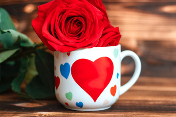 White mug with hearts on a background of wooden boards. Red roses on a top of mug. Attributes for the celebration of Valentine's day