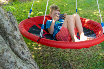 Happy boy on a swing playing phone games outside. Screen time limit - concentrated involved child browsing the internet.