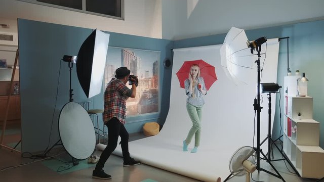 Backstage of the photo shoot: beautiful blonde model posing with red umbrella. Professional equipment studio