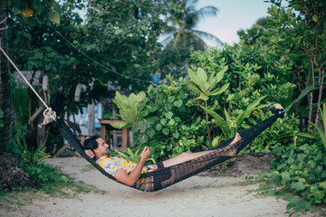 A young man lies in a hammock on a sunny beach with a phone.
