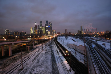 Fototapeta na wymiar Industrial landscape with many railroad tracks going far, cars on the overpass and skyscrapers with power plant pipes on horizon on twilight in winter season