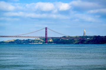 Lisbon, Portugal - Suspension Red Bridge on April 25 across the Tagus River, amid clouds in the blue sky, a statue of Christ on the other side of the river, in the summer afternoon.